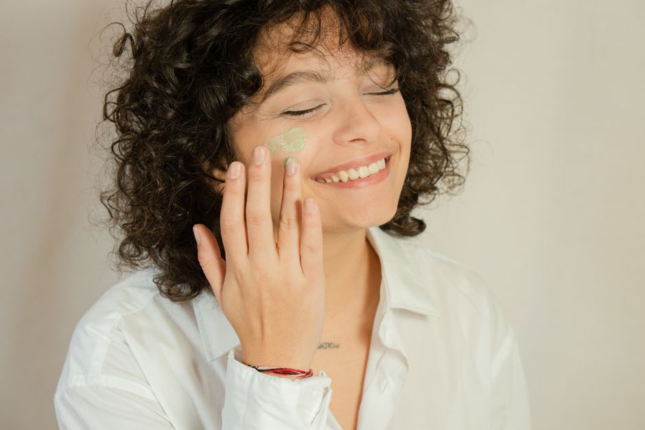 Close-up of a smiling woman applying facial cream with eyes closed.