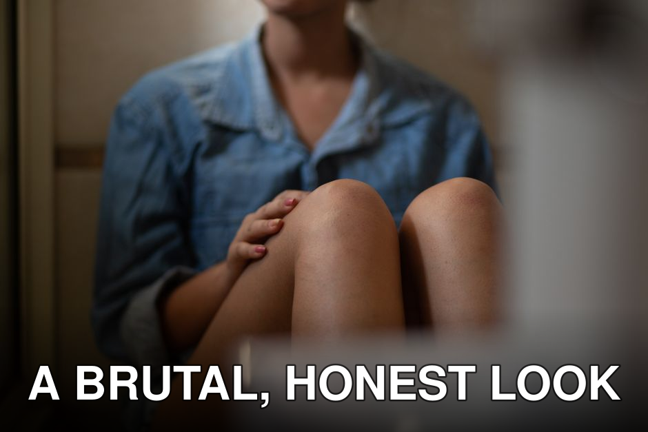 A young woman sits contemplatively indoors wearing a denim jacket, focused on her knees.