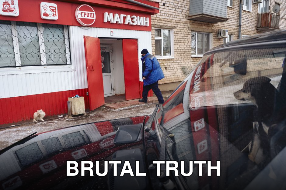 A man in a blue coat enters a red store while a dog watches from a car parked nearby.