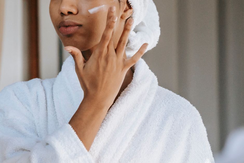 Crop anonymous female in white bathrobe with towel on head applying facial moisturizing cream on face while standing in bathroom