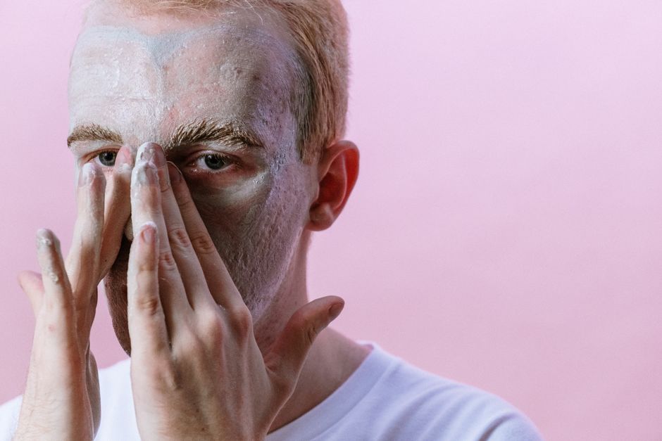 Young man applying clay mask as part of his skincare routine against pink background.