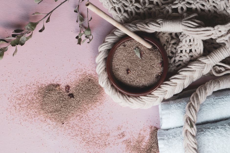 Flat lay of spa essentials including natural scrub, towels, and greenery on a pink surface.