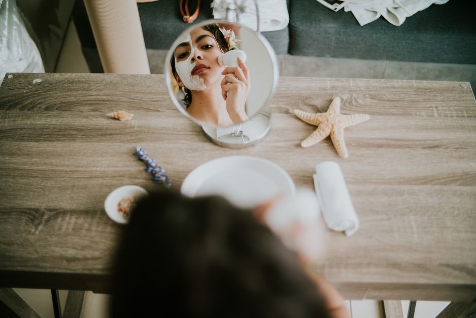 A woman applies a facial mask in front of a mirror with spa elements on a wooden table.
