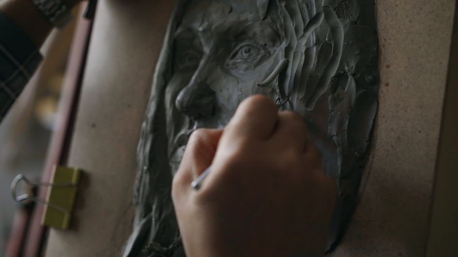 Close-up of a hand sculpting a clay face on an artist's easel indoors.