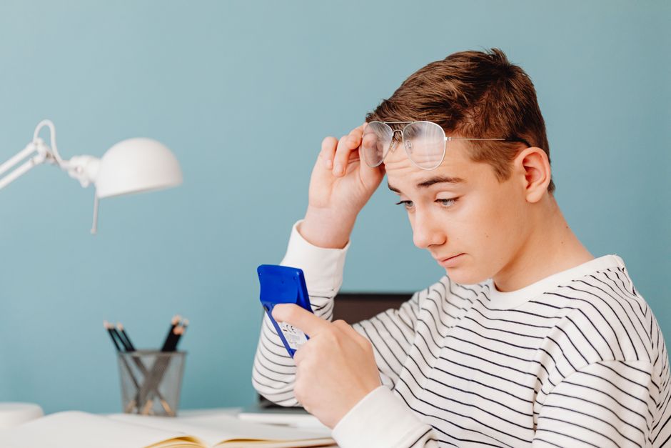 Teenage boy using a blue calculator, adjusting glasses, focused inside a room with a blue wall.
