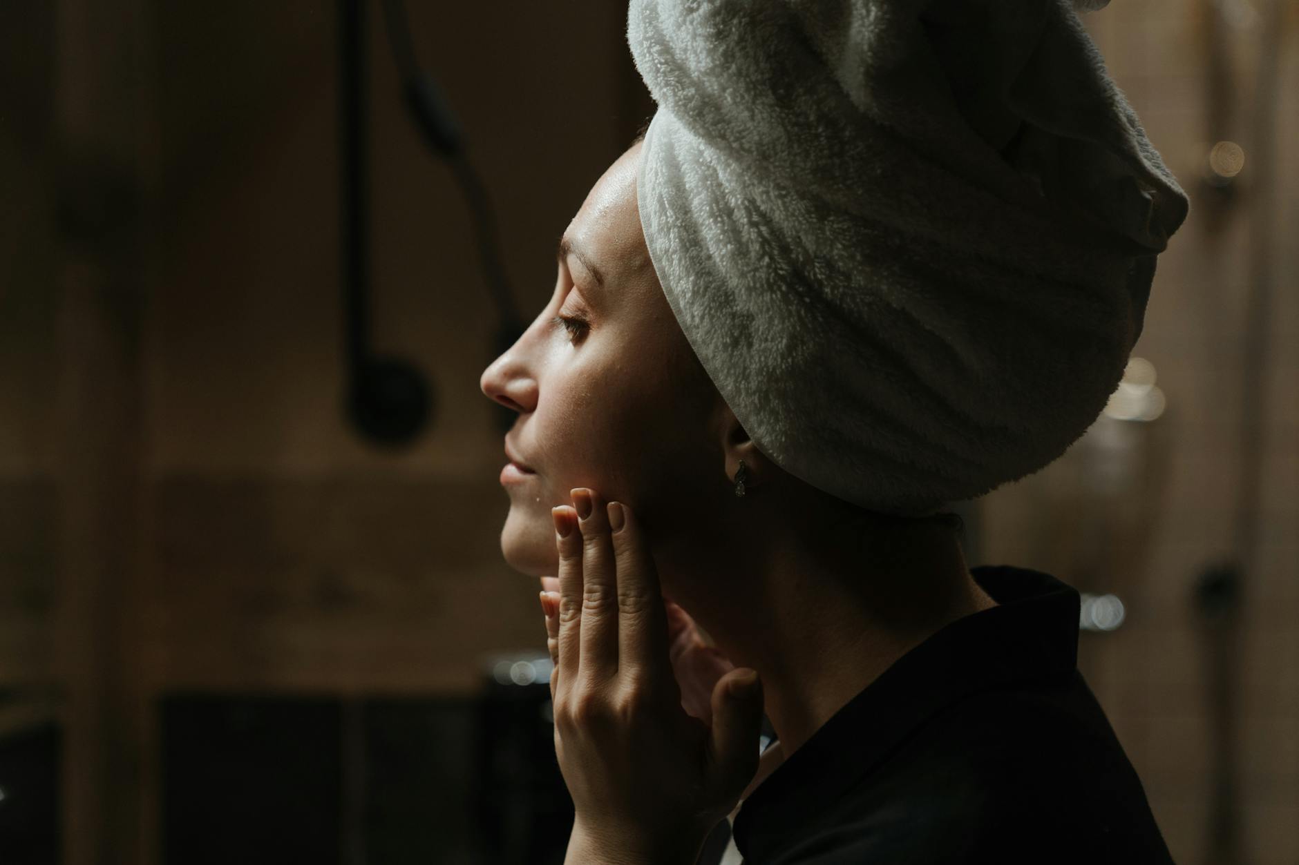 Woman with towel headwrap enjoying a skincare routine in dim lighting for self-care.