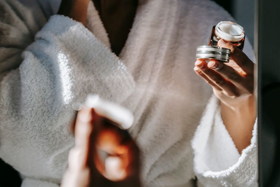 Crop unrecognizable ethnic female with open jar of moisturizing cream reflecting in mirror during daily routine at home