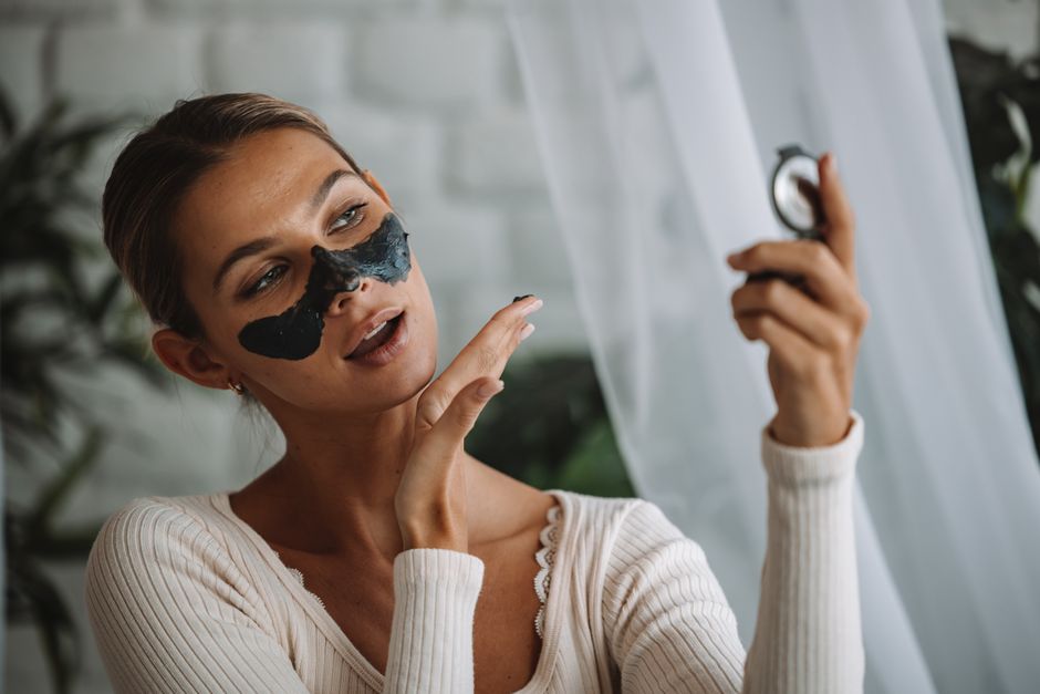 Woman applying black peel-off face mask while looking in a mirror indoors.