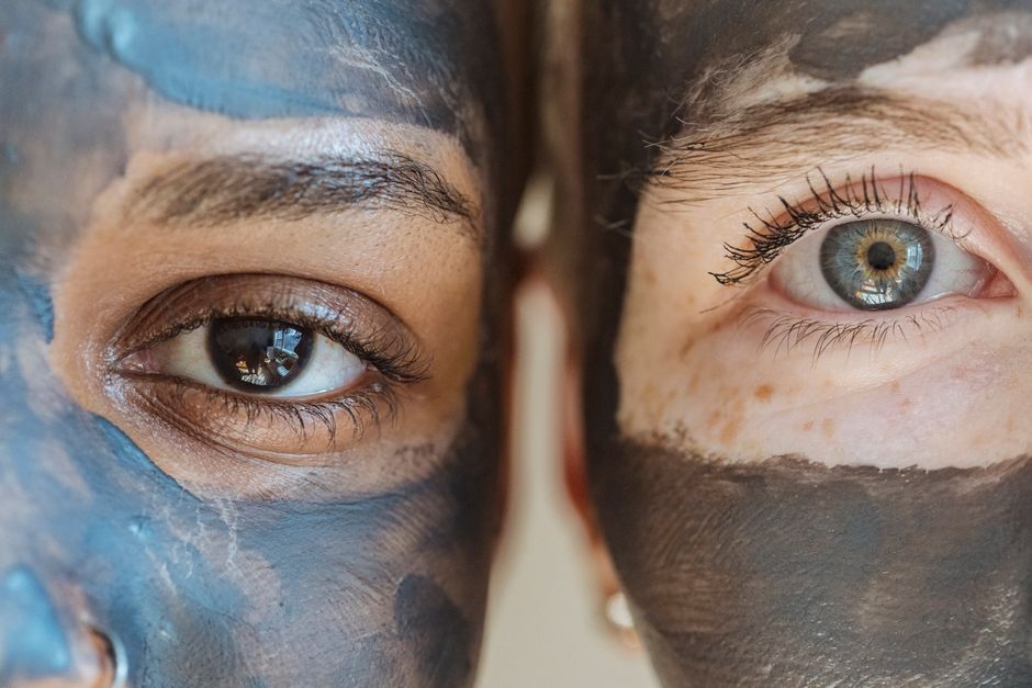 Closeup of diverse crop females with clay mask on faces looking at camera during skincare routine