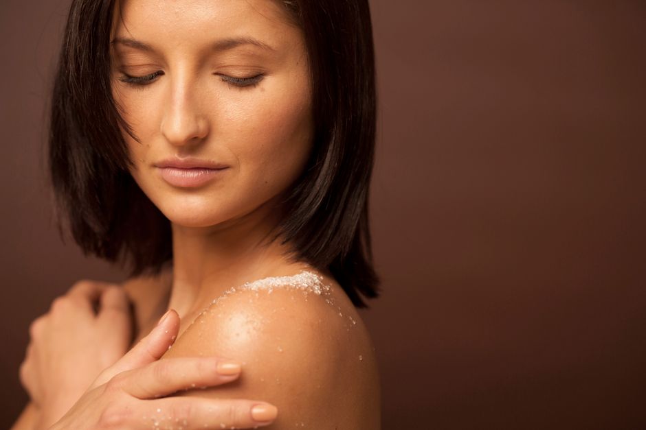 Close-up of a woman enjoying a skincare routine, with exfoliating salt on her shoulder, in a warm studio setting.