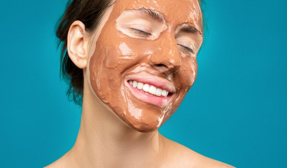 Close-up of a woman smiling with a clay mud face mask, emphasizing skincare and relaxation.