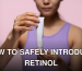 A woman in a purple tank top holds a skincare cream against a pink background in a studio setting.