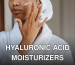 Crop anonymous female in white bathrobe with towel on head applying facial moisturizing cream on face while standing in bathroom