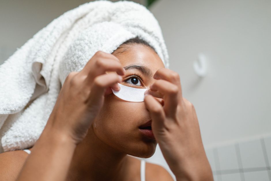 A woman with a towel on her head applies under eye masks during her skincare routine.
