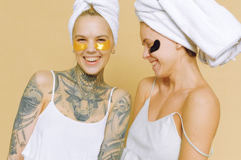 Cheerful young ladies in white outfit and bath towels on head having fun while standing near each other in hydrogel eye patches under eyes