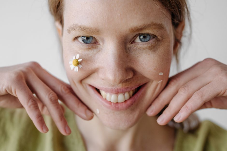 Close-up photo of a smiling woman with a flower on her cheek, photographed indoors.