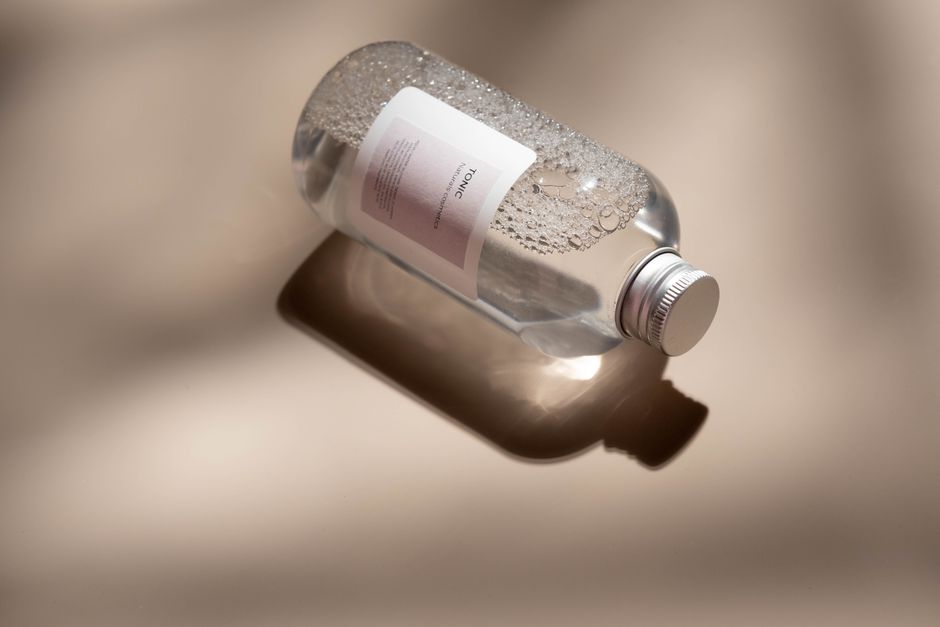 A clear plastic bottle of skincare tonic with bubbles rests on a reflective surface with ample copyspace.