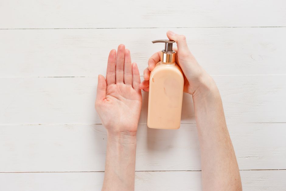 Close-up of a person's hands applying lotion using a pump bottle on a white wooden background.