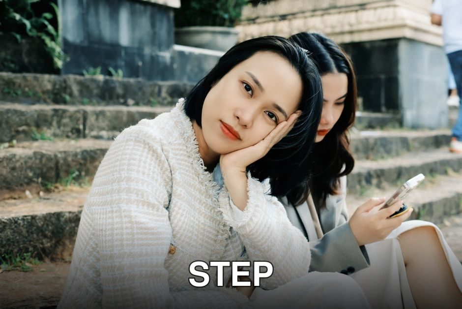 Two women sitting on stone steps, one posing thoughtfully while the other uses her phone.