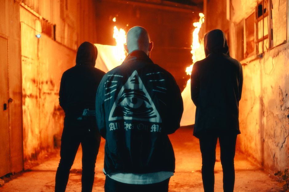 Three hooded figures stand in an urban alley, surrounded by fire at night. Mystery abounds.