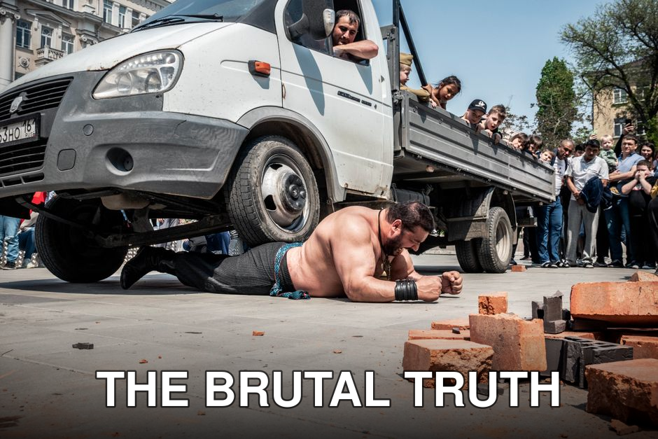 A crowd watches a strongman perform a daring feat under a truck on a busy street.