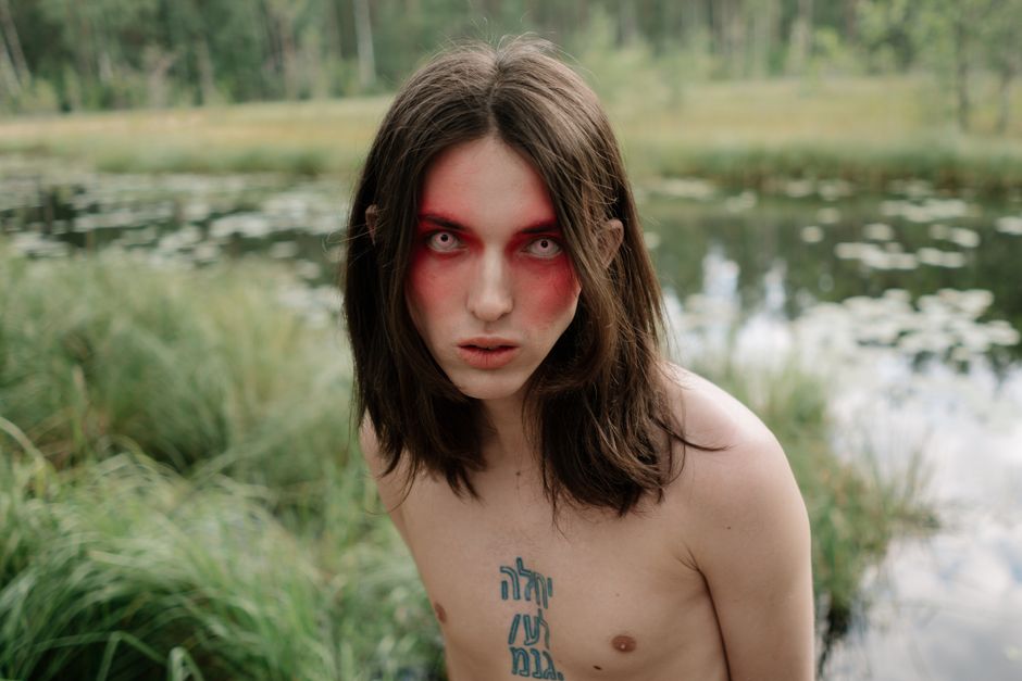 Eerie portrait of a person with red makeup and tattoos by a pond.