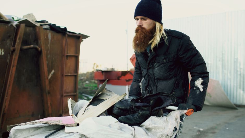 Bearded man in winter clothes sorting items from a dumpster outdoors, symbolic of urban survival.