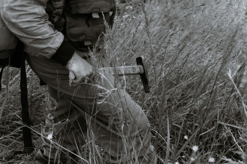 Black and white side view of crop unrecognizable male in camouflage outfit carrying backpack and hammer while walking through grassy meadow