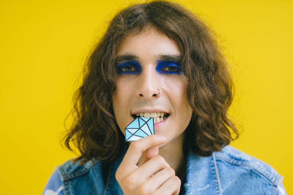 Portrait of a man with striking blue eye makeup holding a geometric shape against a bright yellow backdrop.