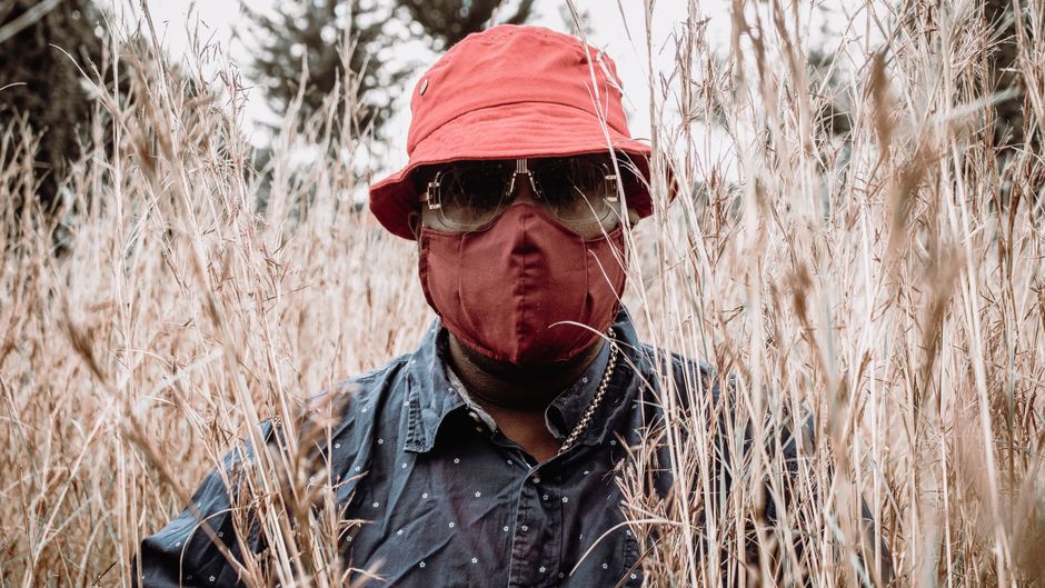 A fashionable man in a red bucket hat and face mask stands amidst tall grass.