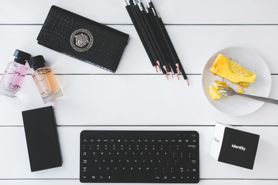 Flat lay of a modern workspace with keyboard, perfume, cake slice, and stationery on white desk.