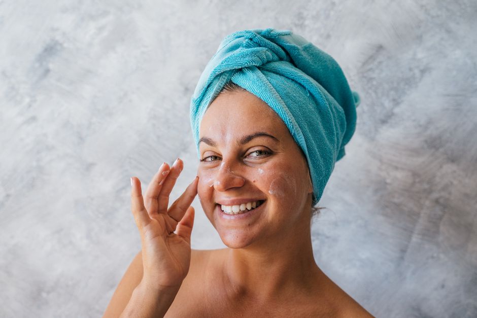 Happy woman with towel on head applies moisturizer, showcasing skincare routine.