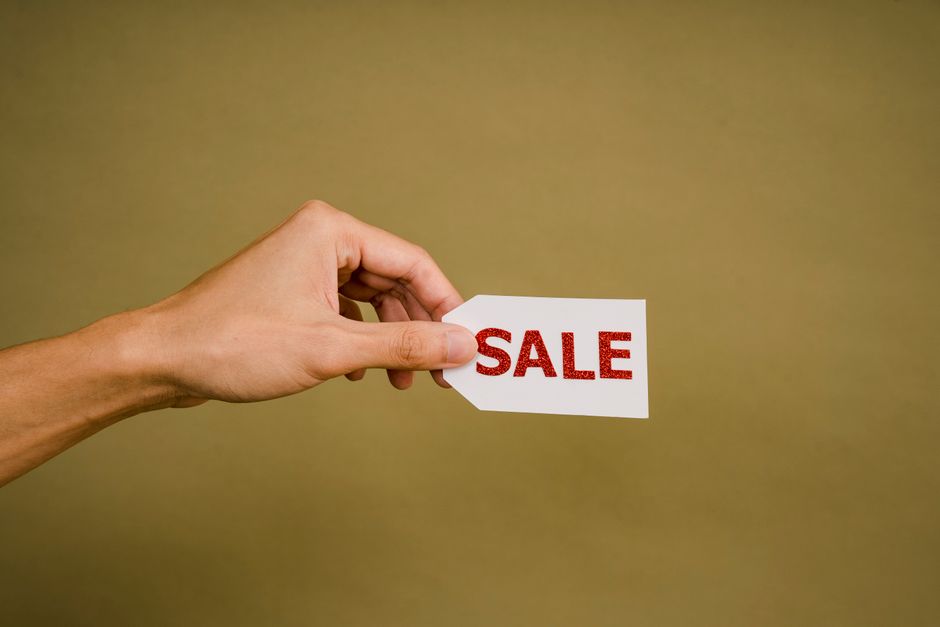 Crop anonymous person showing small white paper tag with inscription Sale against brown wall