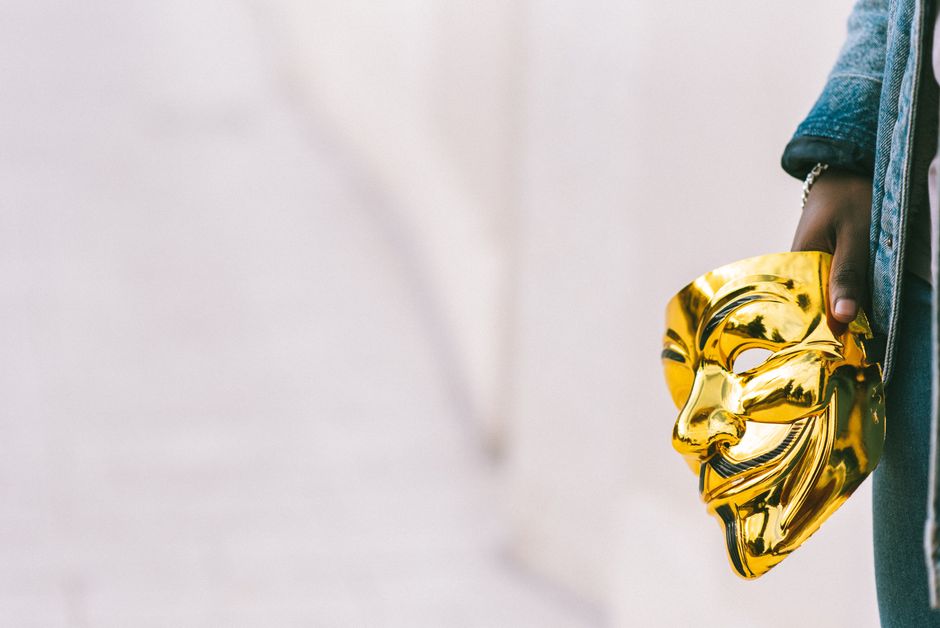 Crop unrecognizable African American person showing golden Anonymous mask in hand against light background
