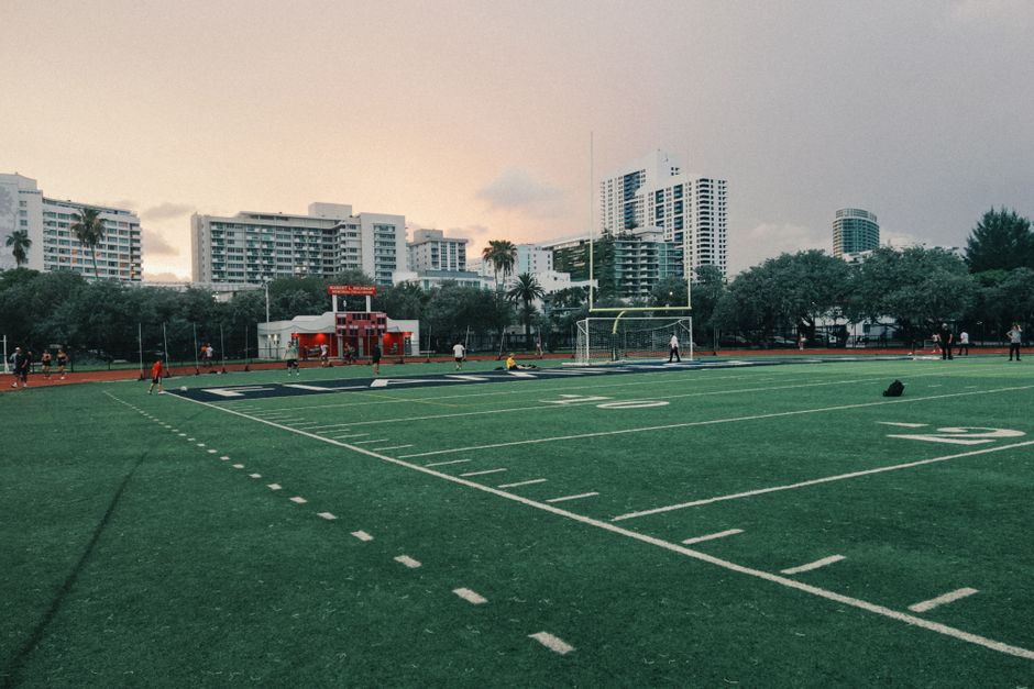 A football field in an urban setting at sunset, surrounded by high-rise buildings.