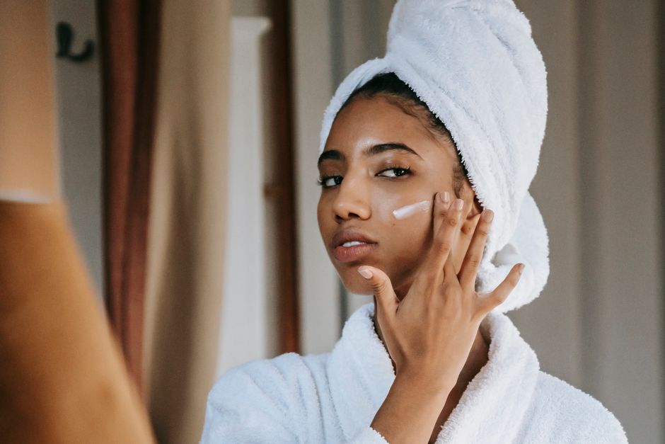 Young attentive ethnic female in bathrobe applying smear of moisturizing cream on face while looking in mirror at home