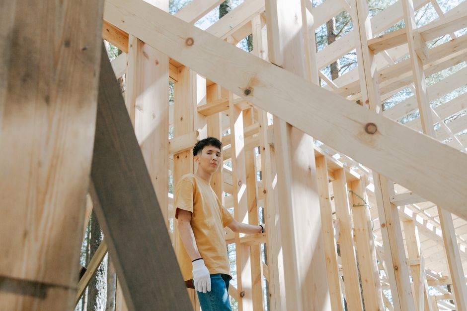 Man in brown shirt and gloves building a wooden structure in sunlight.