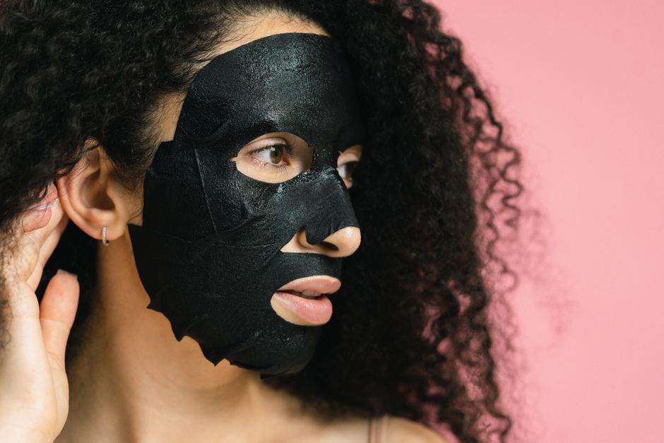 Close-up of a woman with black facial mask and curly hair, set against a pink backdrop.