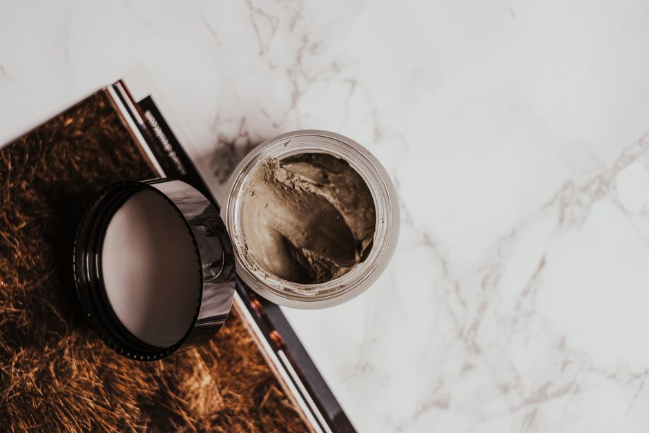 High-angle view of a clay mask in a glass jar on a marble surface with a magazine.