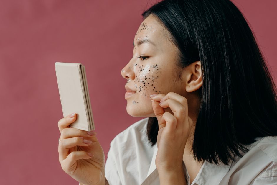 A young woman applies a glitter facial mask while looking into a compact mirror, set against a pink background.