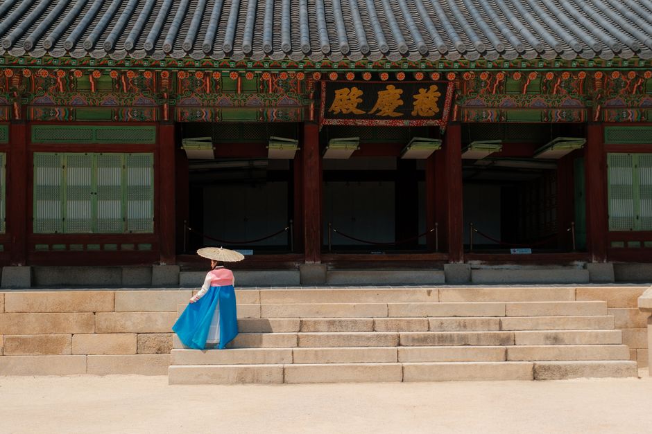 A person wearing Korean Hanbok stands in front of a traditional palace in Seoul, South Korea.