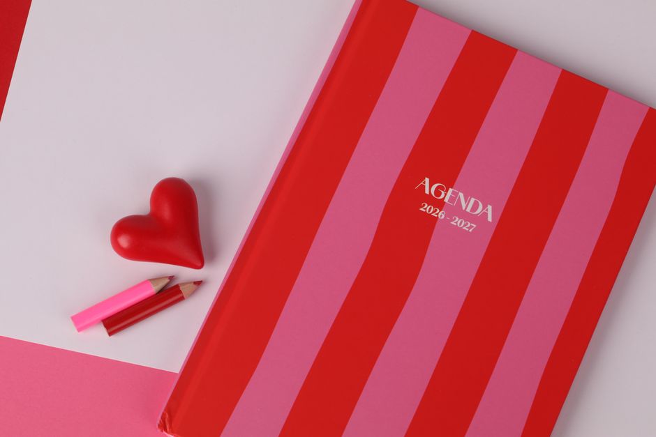 Flat lay of a red and pink striped agenda, heart-shaped object, and colored pencils, ideal for planning themes.