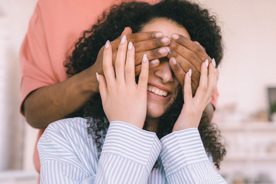A smiling woman with curly hair playfully covers her eyes in a joyful moment indoors.