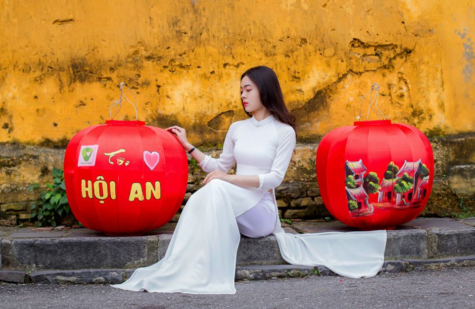 Woman in white Áo Dài sits by red lanterns in Hội An, Vietnam.